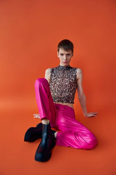 Queer persona en leopardo imprimir sin mangas superior y rosa pantalones sentado en naranja telón de fondo, moda - foto de stock