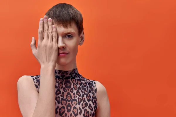 Queer person in leopard print sleeveless top covering half of face on orange backdrop, short hair — Stock Photo