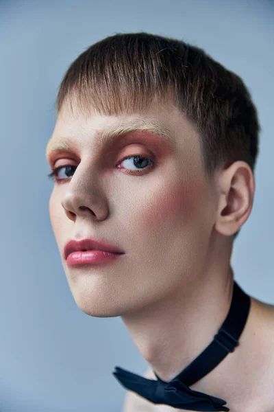 Modelo queer con maquillaje mirando a la cámara, fondo gris, moda, pajarita, andrógino, retrato - foto de stock