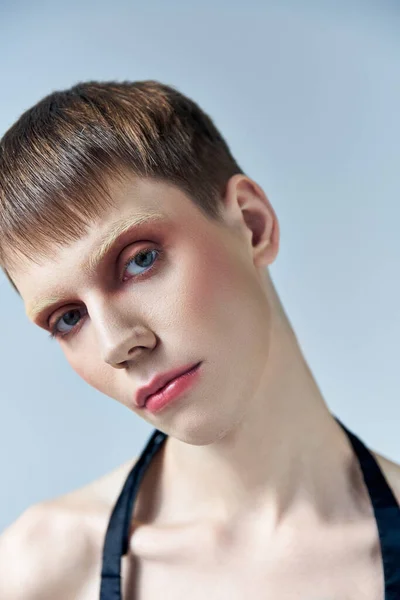 Retrato, persona andrógina con maquillaje mirando a la cámara en gris, belleza y rostro, queer - foto de stock