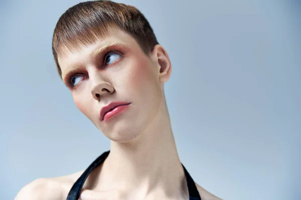 Retrato, modelo andrógino con maquillaje mirando hacia otro lado sobre fondo gris, belleza y rostro, queer - foto de stock