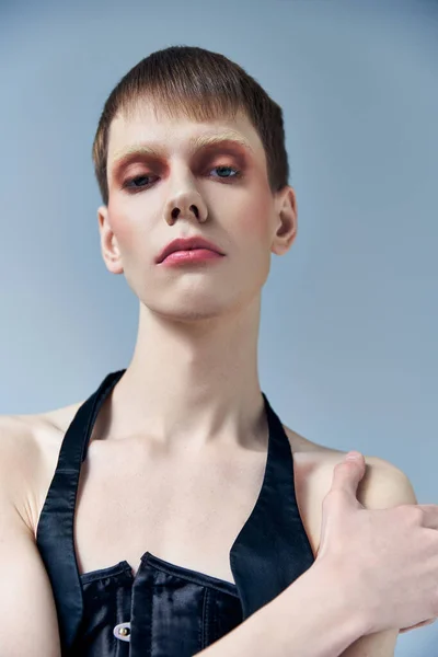 Retrato, modelo queer con maquillaje mirando a la cámara, posando sobre gris, belleza y rostro, andrógino - foto de stock