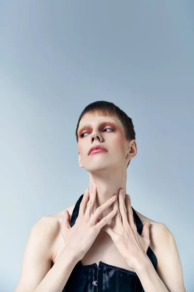 Retrato, modelo queer com maquiagem tocando pescoço e posando em cinza, beleza e visagem, andrógino — Fotografia de Stock