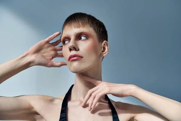 Persona queer con maquillaje mirando hacia otro lado y posando sobre fondo gris, belleza, rostro, andrógino - foto de stock