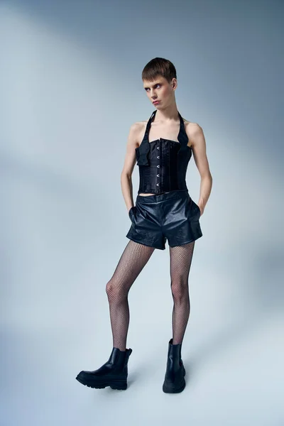 Queer persona en corsé negro y pantalones cortos posando con las manos en bolsillos sobre fondo gris, andrógino - foto de stock