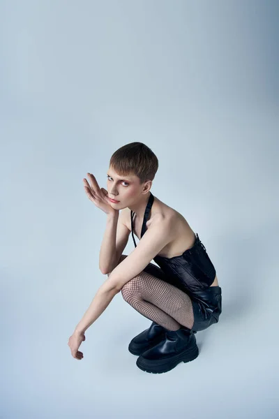 Modelo en corsé negro y pantalones cortos sentados sobre fondo gris, persona andrógina, moda queer - foto de stock