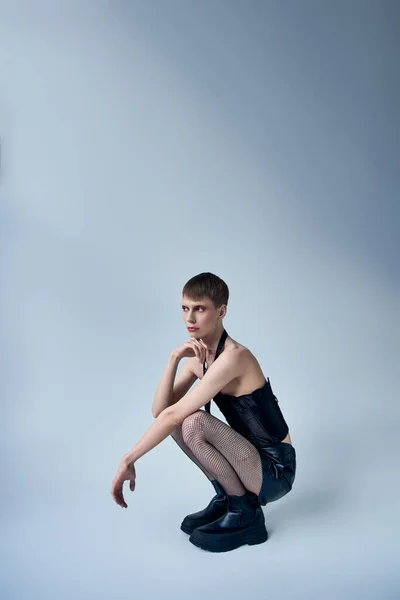 Modelo queer en corsé negro y medias de rejilla sentado sobre fondo gris, persona andrógina, moda - foto de stock