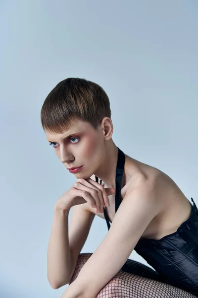Modèle queer en corset noir et collants résille assis sur fond gris, style de personne androgyne — Photo de stock