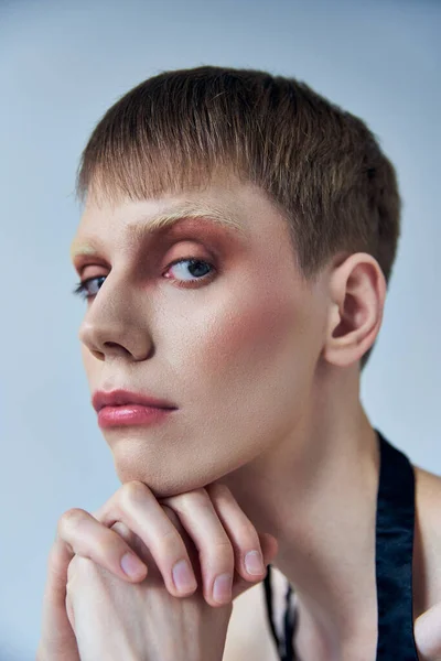 Modelo queer con maquillaje mirando a la cámara en el fondo gris, persona andrógina, retrato, identidad - foto de stock