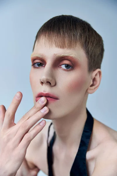 Queer person with makeup on grey backdrop, androgynous person, touching lip, look at camera — Stock Photo
