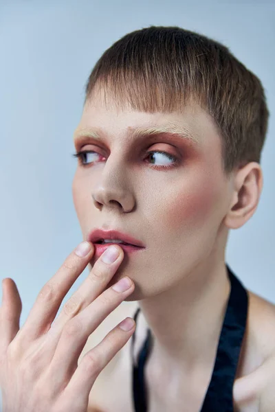Queere Person mit Make-up, die vor grauem Hintergrund wegschaut, androgyne, anrührende Lippe, Selbstausdruck — Stockfoto