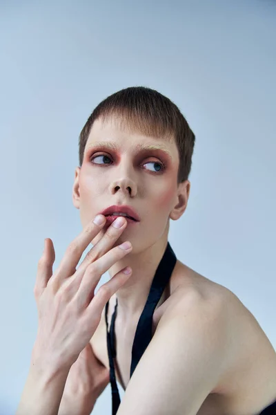 Modelo queer con maquillaje mirando hacia otro lado en el fondo gris, andrógino, labio conmovedor, auto expresión - foto de stock