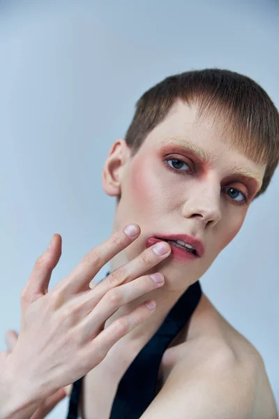 Queer person with makeup looking at camera on grey, androgynous, touching lip, self expression — Stock Photo