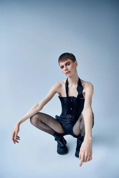 Modelo queer en corsé negro, botas, medias de rejilla sentado en gris, estilo de persona andrógina - foto de stock