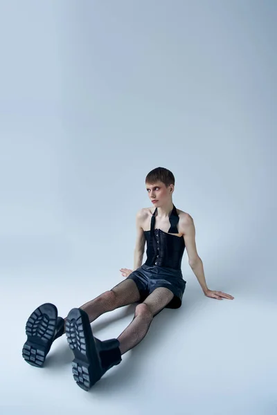Persona queer en corsé negro, pantalones cortos, medias de red de pesca sentado en gris, modelo andrógino, estilo lgbt - foto de stock