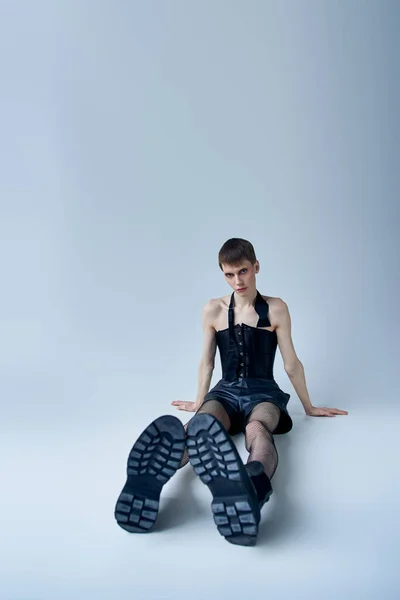 Modelo queer en corsé negro y medias de rejilla sentado en gris, persona andrógina, lgbt moda - foto de stock