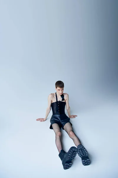 Queer person in black corset and fishnet tights sitting on grey, androgynous model, lgbt fashion — Stock Photo