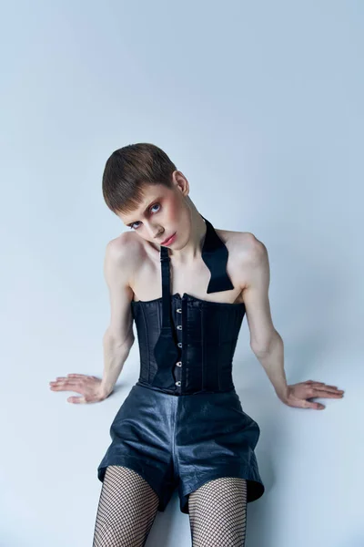 Queer person in black corset and fishnet tights sitting on grey, lgbt, androgynous model, fashion — Stock Photo
