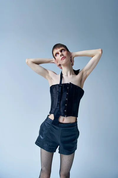 Modelo andrógino en corsé negro y pantalones cortos de pie sobre fondo gris, lgbt, moda queer - foto de stock