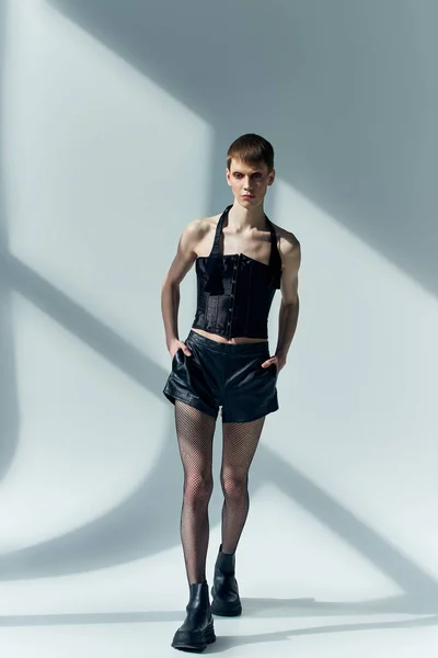Modello androgino in corsetto e pantaloncini neri in posa su grigio, lgbt, moda queer, mani in tasca — Foto stock
