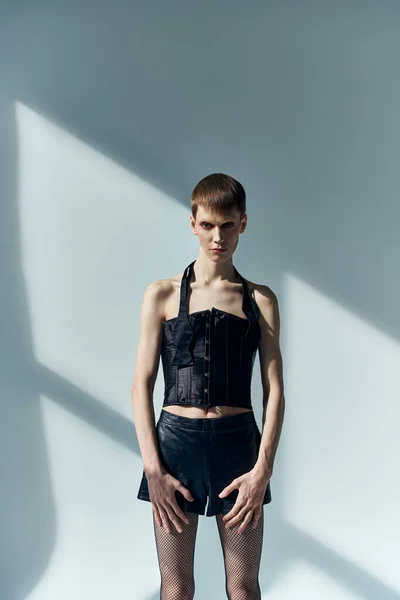 Androgynous model in corset and shorts posing on grey backdrop with shadows, lgbt, queer fashion — Stock Photo