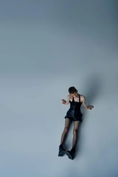 Modèle androgyne en collants corset et résille assis sur fond gris, style queer, angle élevé — Photo de stock