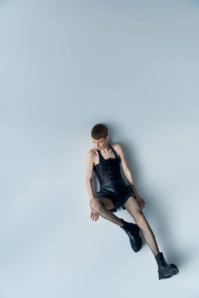 Queeres Modell in Korsett und Netzstrumpfhose auf grauem, lgbt, androgynem Stil sitzend, Blick in den hohen Winkel — Stockfoto