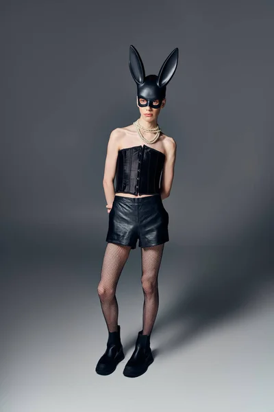 Modelo andrógino en corsé negro posando en bdsm conejito máscara sobre fondo gris, moda queer, lgbt - foto de stock