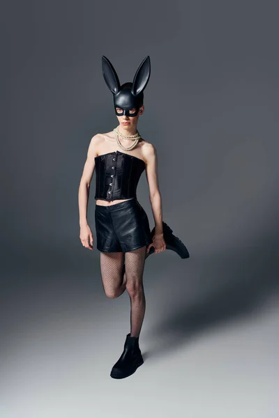 Modello androgino in corsetto posa in bdsm maschera coniglietto su grigio, moda queer, provocatorio — Foto stock