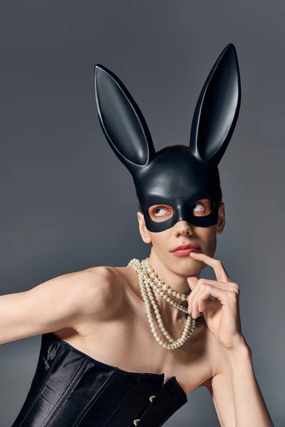 Nachdenkliches Model im Korsett posiert in bdsm Hasenmaske auf grau, sitzt, schaut weg, queere Mode — Stockfoto