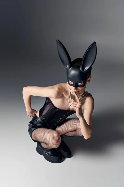 Provocatorio queer persona in corsetto posa in bdsm coniglietto maschera su grigio, seduta, tagliente moda — Foto stock