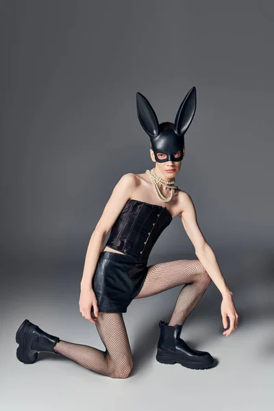 Olhar ousado, pessoa provocante em espartilho posando em bdsm máscara de coelho em cinza, moda queer, estilo — Fotografia de Stock