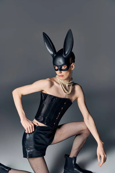 Смелый стиль, провокационный человек в корсете, позирующий в маске кролика на серой, причудливой моде, отворачивается — стоковое фото
