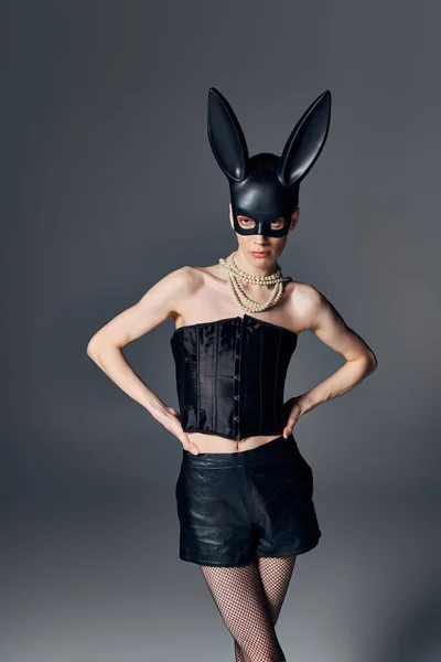 Moda, genderqueer persona in corsetto posa in bdsm maschera coniglietto su grigio, stile queer, mani sui fianchi — Foto stock