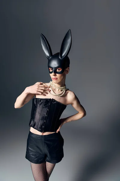 Moda, genere fluido persona in corsetto posa in bdsm maschera coniglietto su grigio, stile queer, mano sul fianco — Foto stock