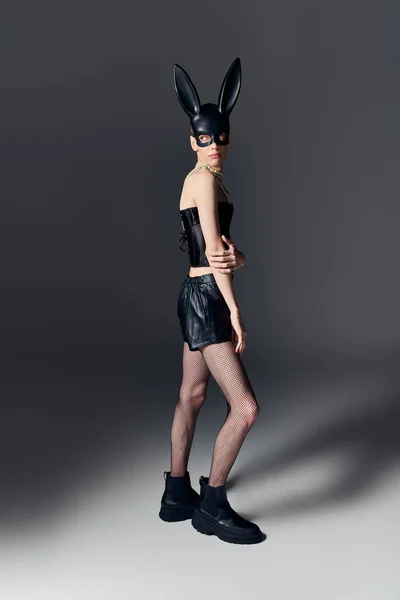 Gender fluid person in bold outfit posing in bdsm bunny mask on grey, queer fashion, full length — Stock Photo