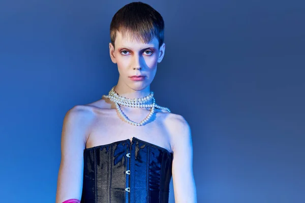 Queer personne en corset noir et collier de perles posant sur fond bleu, mode agitée, non binaire — Photo de stock