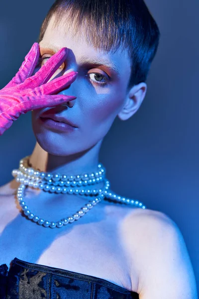 Modèle queer en collier de perles couvrant le visage avec la main en gant rose sur fond bleu, mode audacieuse — Photo de stock