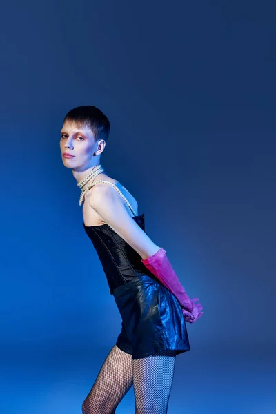 Nonbinary person in corset and black shorts posing on blue backdrop, queer model in pink gloves — Stock Photo