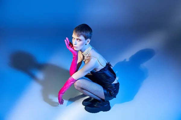 Persona no binaria en corsé y pantalones cortos negros sentados sobre fondo azul, modelo queer en guantes rosados - foto de stock