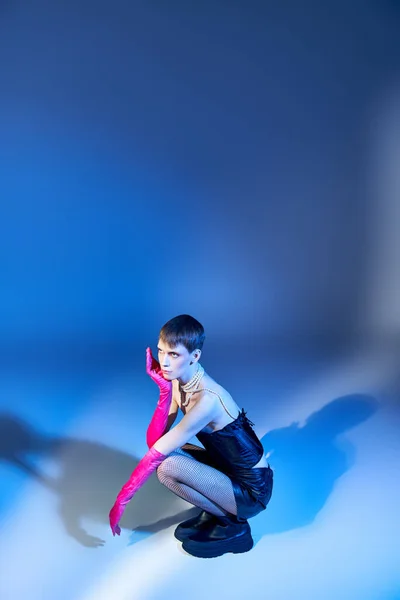 Queer en corset et short noir assis sur fond bleu, modèle non binaire en gants roses — Photo de stock