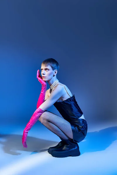 Modello queer in corsetto nero e pantaloncini seduti su sfondo blu, persona non binaria in guanti rosa — Foto stock