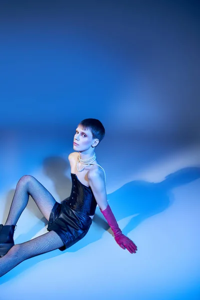 Jeune modèle queer en corset et short assis sur fond bleu, personne non binaire en gants roses — Photo de stock