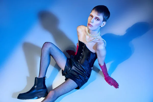 Modèle queer en corset et short assis sur fond bleu, personne non binaire en gants roses, mode — Photo de stock
