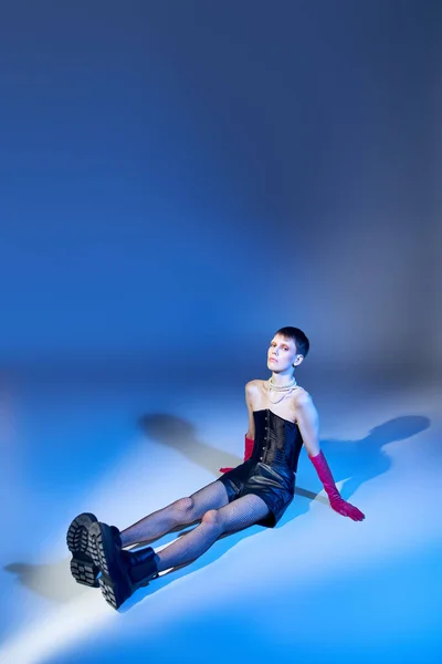 Modèle queer en corset et bottes assis sur fond bleu, personne non binaire en gants roses, mode — Photo de stock