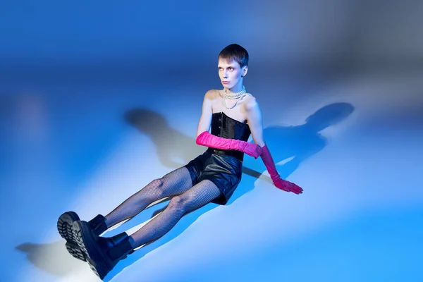 Modell in Korsett und Stiefeln auf blauem Hintergrund sitzend, nichtbinäre Person in rosa Handschuhen, queere Mode — Stockfoto