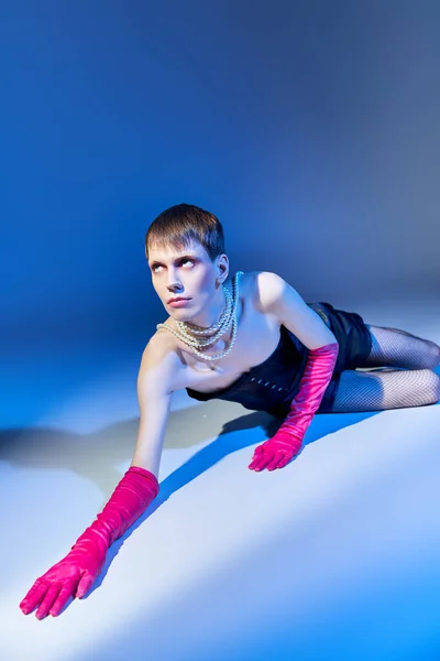 Queer model in bold outfit and pink gloves posing on blue backdrop, shorts, nonbinary, look up — Stock Photo