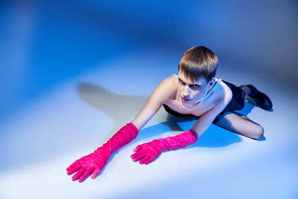 Modelo queer en traje negrita y guantes de color rosa posando sobre fondo azul, no binario y extravagante - foto de stock