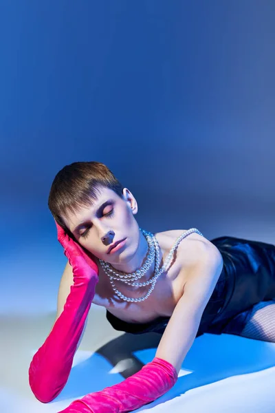 Verträumtes queeres Model in fettem Outfit und rosa Handschuhen posiert auf blauem Hintergrund, nichtbinär, geschlossene Augen — Stockfoto