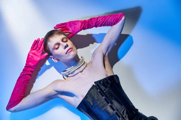 Vista superior, modelo queer en corsé y guantes de color rosa acostado sobre fondo azul, no binario, ojos cerrados - foto de stock